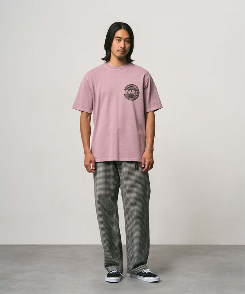Model wearing a lilac pigment colored Gramicci Icon Tee Pigment Dyed with a graphic and grey pants on a light-tone background.