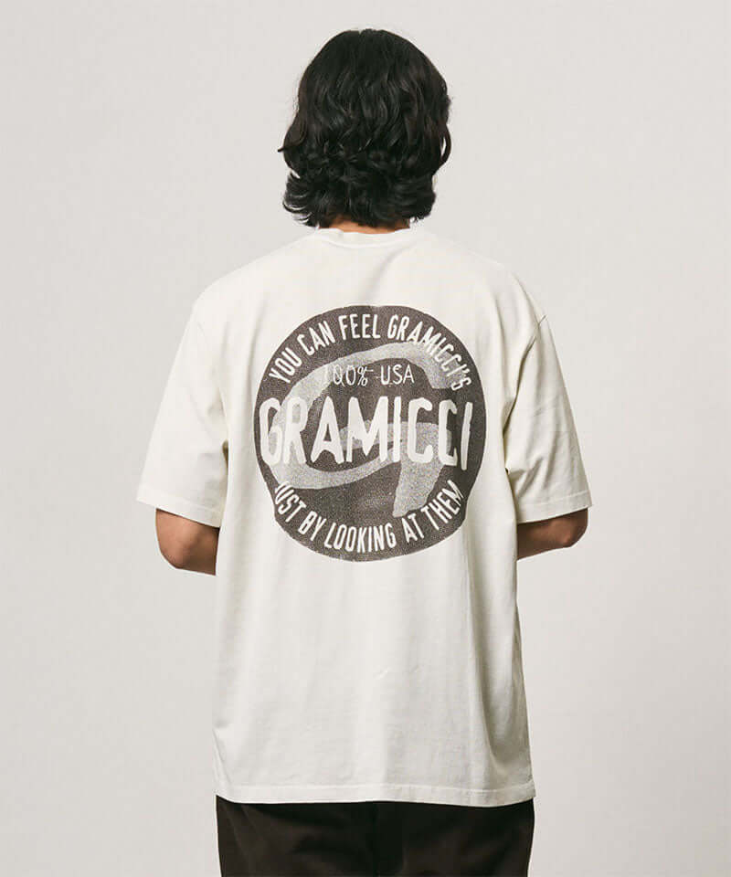 Back view of a model wearing a Gramicci natural pigment colored Icon Tee Pigment Dyed with a graphic against a light-tone background.