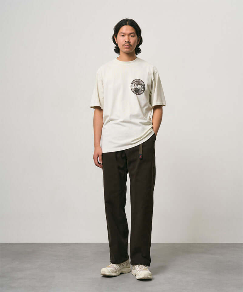 Model wearing a natural pigment colored Gramicci Icon Tee Pigment Dyed with a graphic and dark brown pants on a light-tone background.