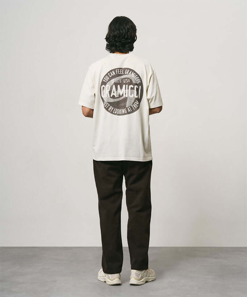 Back view of a model wearing a natural pigment colored Gramicci Icon Tee Pigment Dyed with a graphic and dark brown pants on a light-tone background.