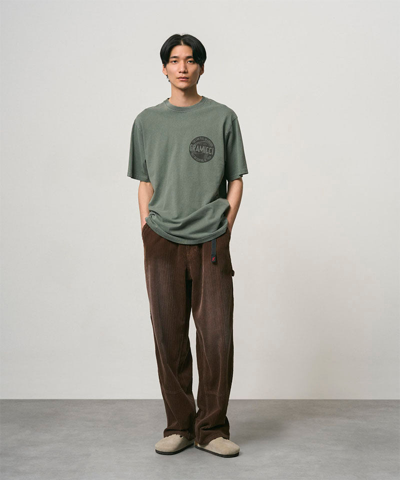 Model wearing a sage pigment colored Gramicci Icon Tee Pigment Dyed with a graphic and dark brown pants on a light-tone background.