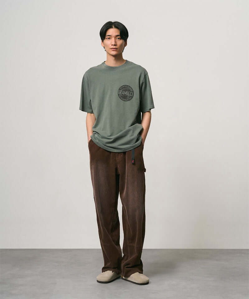 Model wearing a sage pigment colored Gramicci Icon Tee Pigment Dyed with a graphic and dark brown pants on a light-tone background.