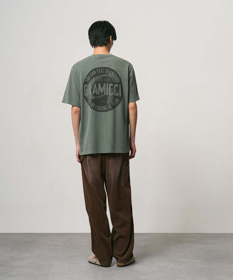 Back view of a model wearing a sage pigment colored Gramicci Icon Tee Pigment Dyed with a graphic and dark brown pants on a light-tone background.