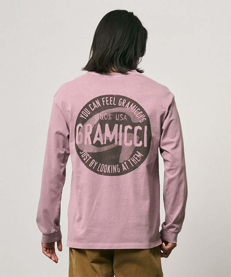 Back view of a model wearing a Gramicci lilac pigment colored Icon Long Sleeve Tee Pigment Dyed with a graphic against a light-tone background.