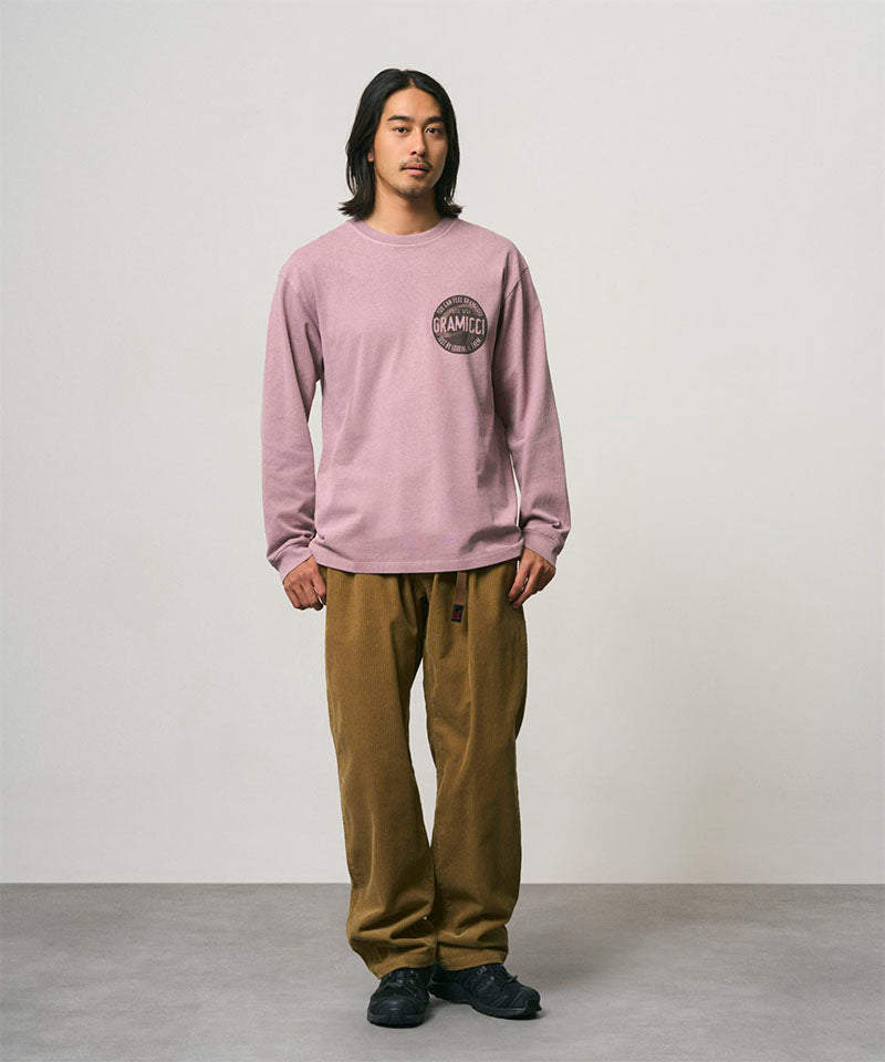 Model wearing a lilac pigment colored Gramicci Icon Long Sleeve Tee Pigment Dyed with a graphic and olive brown pants on a light-tone background.