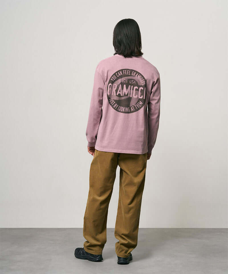 Back view of a model wearing a lilac pigment colored Gramicci Icon Long Sleeve Tee Pigment Dyed with a graphic and olive brown pants on a light-tone background.