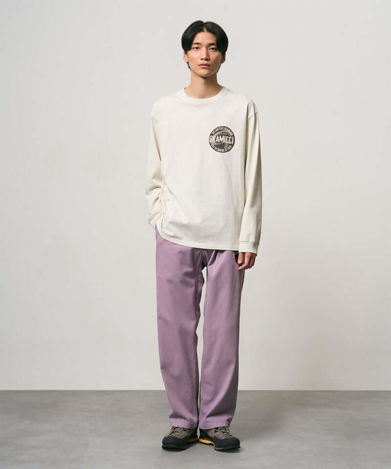 Model wearing a natural pigment colored Gramicci Icon Long Sleeve Tee Pigment Dyed with a graphic and lavender pants on a light-tone background.