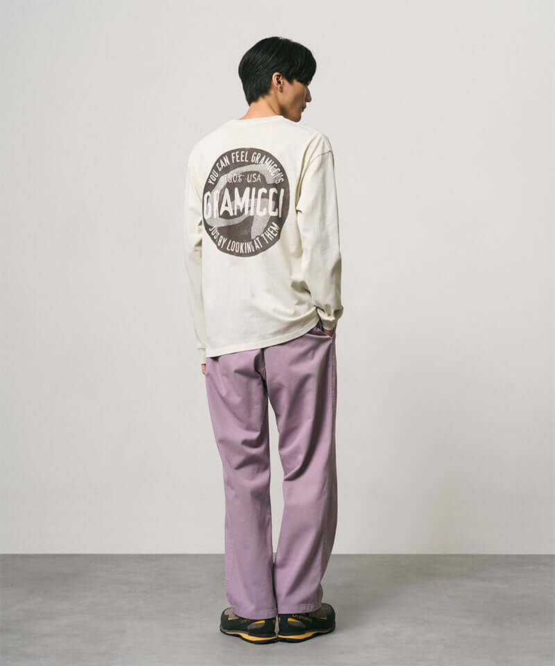 Back view of a model wearing a natural pigment colored Gramicci Icon Long Sleeve Tee Pigment Dyed with a graphic and lavender pants on a light-tone background.