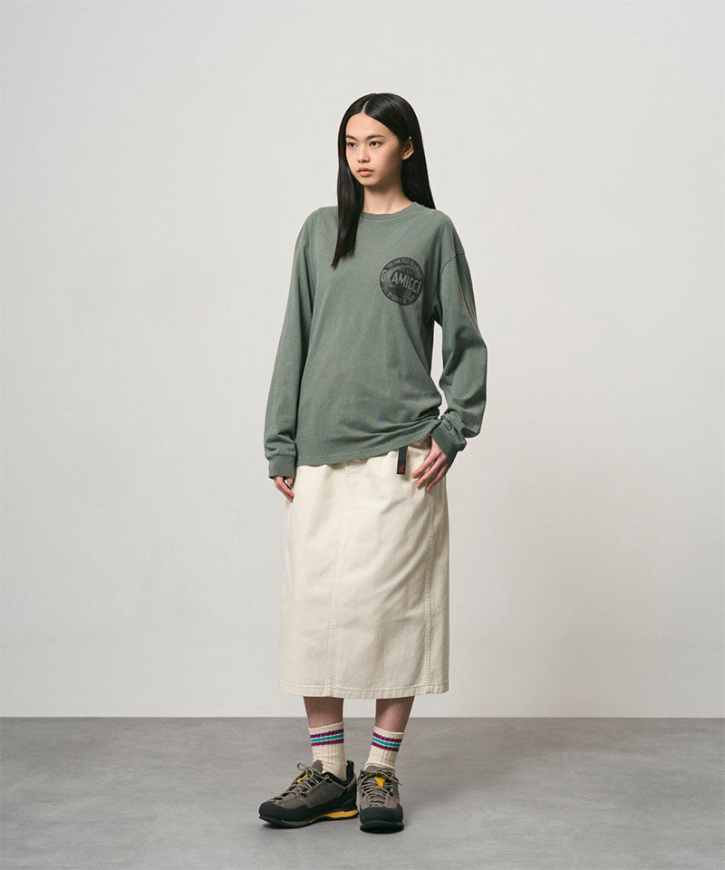 Model wearing a sage pigment colored Gramicci Icon Long Sleeve Tee Pigment Dyed with a graphic and creme colored skirt on a light-tone background.