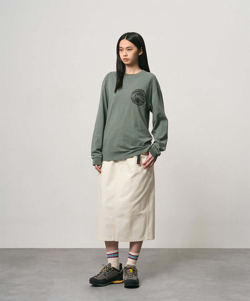 Model wearing a sage pigment colored Gramicci Icon Long Sleeve Tee Pigment Dyed with a graphic and creme colored skirt on a light-tone background.