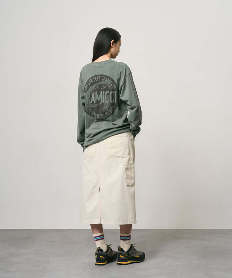 Back view of a model wearing a sage pigment colored Gramicci Icon Long Sleeve Tee Pigment Dyed with a graphic and creme colored skirt on a light-tone background.