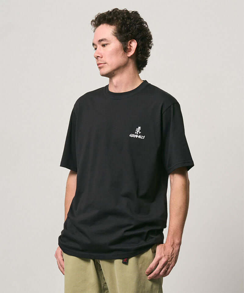 Model wearing a Gramicci black One Point Logo Tee with a graphic against a light-tone background.