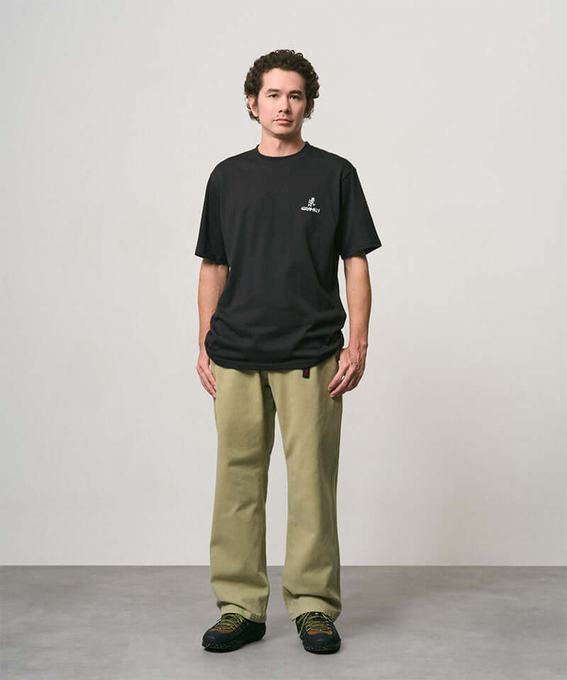Model wearing a black Gramicci One Point Logo Tee with a graphic and light olive pants on a light-tone background.