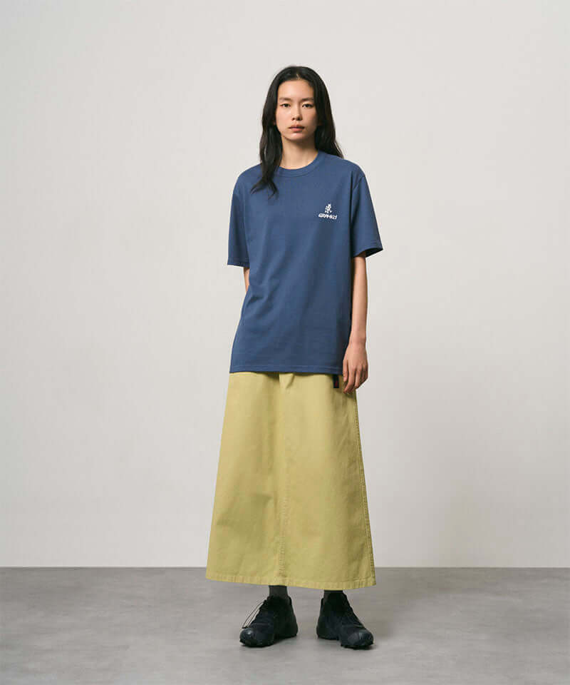Model wearing a dusky blue Gramicci One Point Logo Tee with a graphic and light olive skirt on a light-tone background.