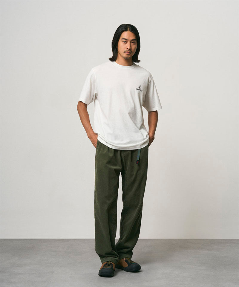 Model wearing a white Gramicci One Point Logo Tee with a graphic and olive pants on a light-tone background.