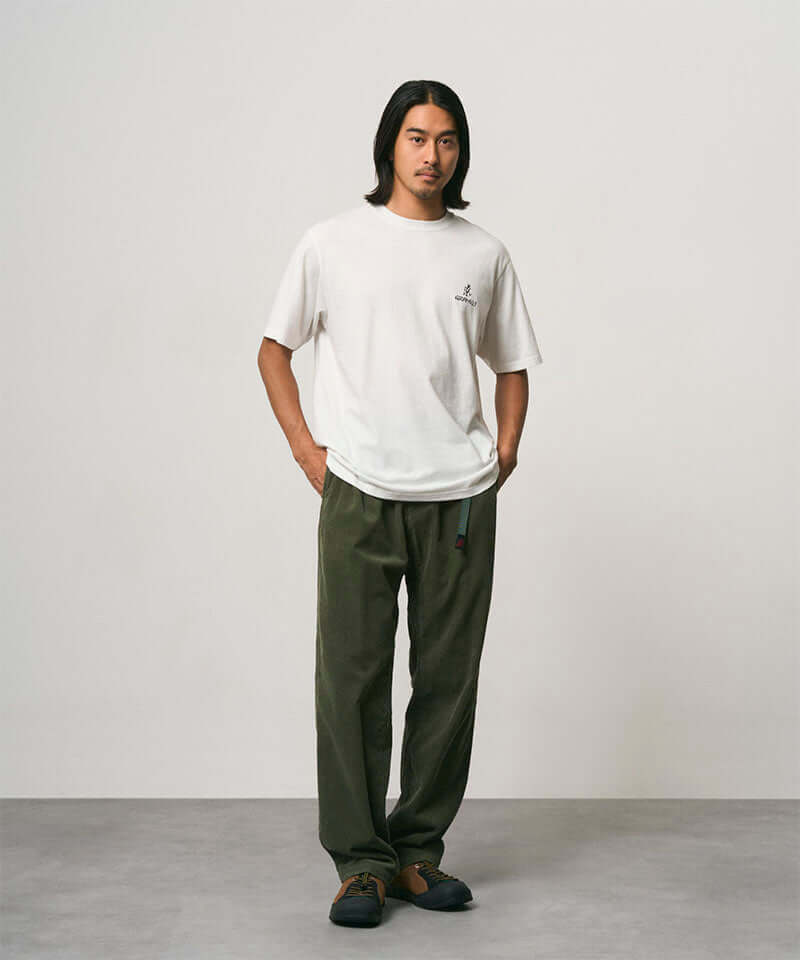 Model wearing a white Gramicci One Point Logo Tee with a graphic and olive pants on a light-tone background.