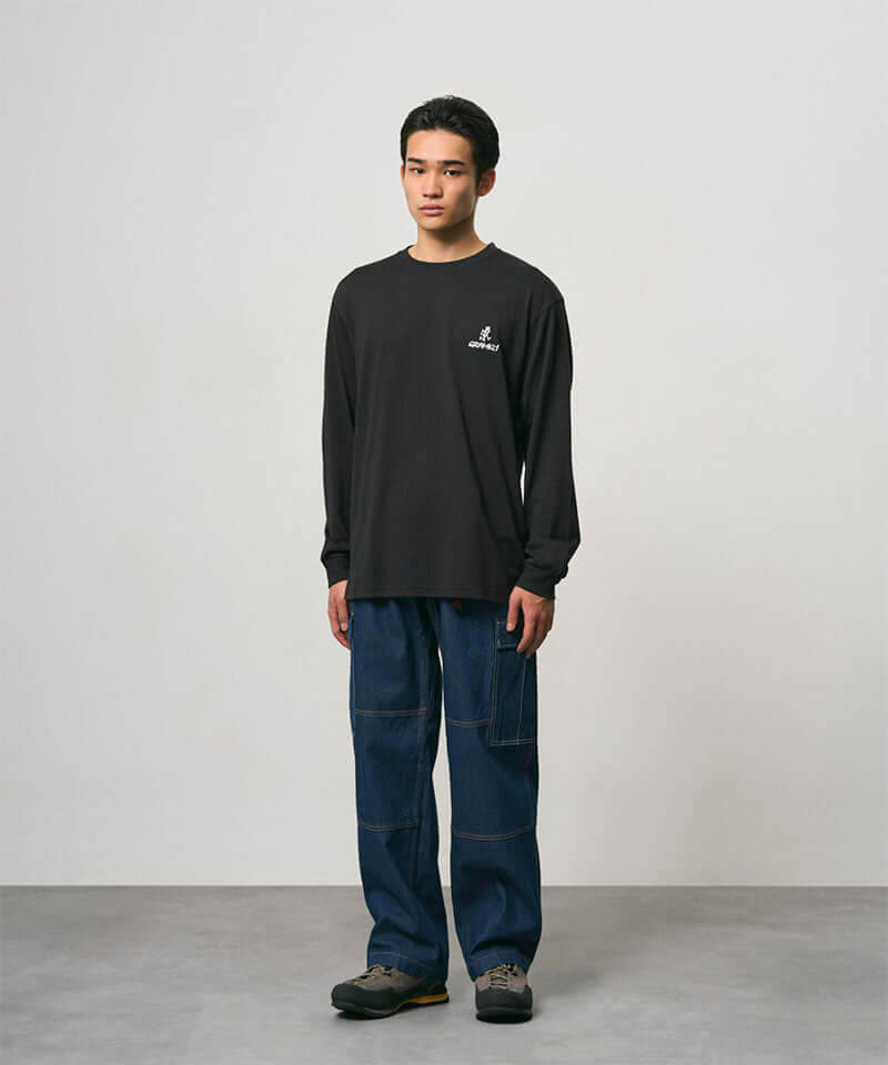 Model wearing a black Gramicci One Point Logo Long Sleeve Tee with a graphic and blue pants on a light-tone background.