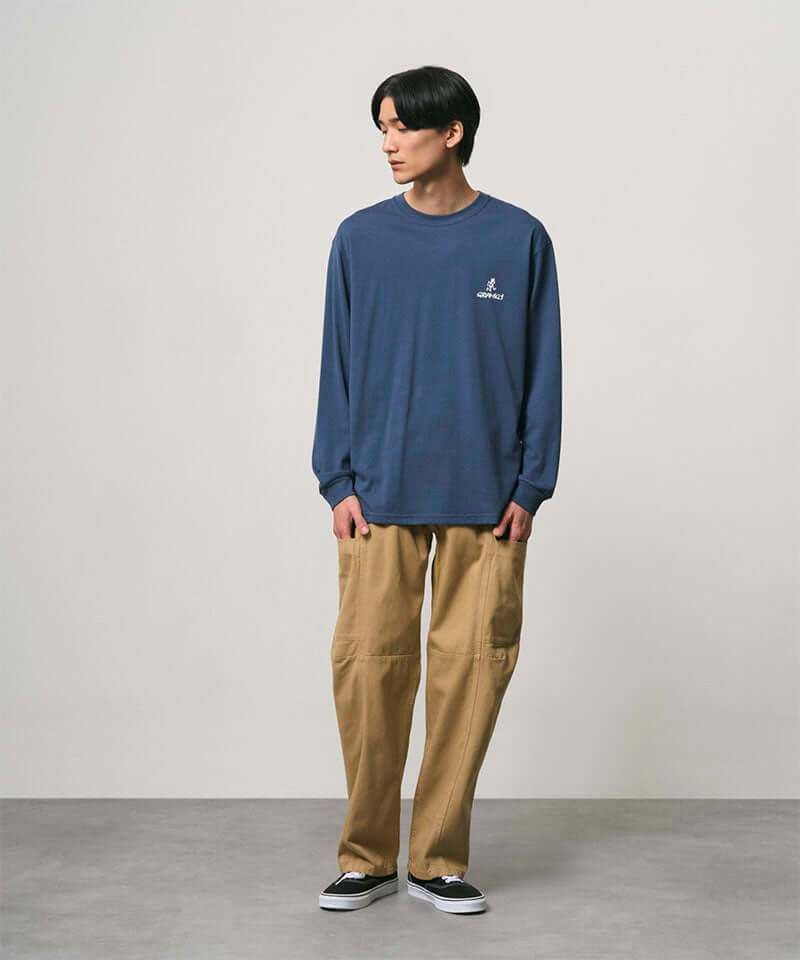Model wearing a dusky blue Gramicci One Point Logo Long Sleeve Tee with a graphic and beige pants on a light-tone background.