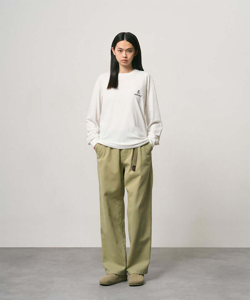 Model wearing a white Gramicci One Point Logo Long Sleeve Tee with a graphic and light olive pants on a light-tone background.