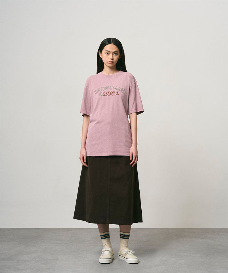 Model wearing a lillac pigment colored Gramicci Livin' Tee Pigment Dyed with a Livin' Under A Rock graphic and black skirt on a light-tone background.