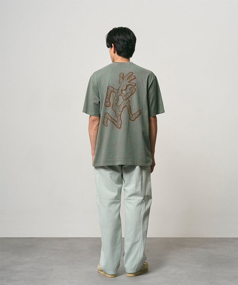 Back view of a model wearing a sage pigment colored Gramicci Livin' Tee Pigment Dyed with a running man graphic and light colored pants on a light-tone background.
