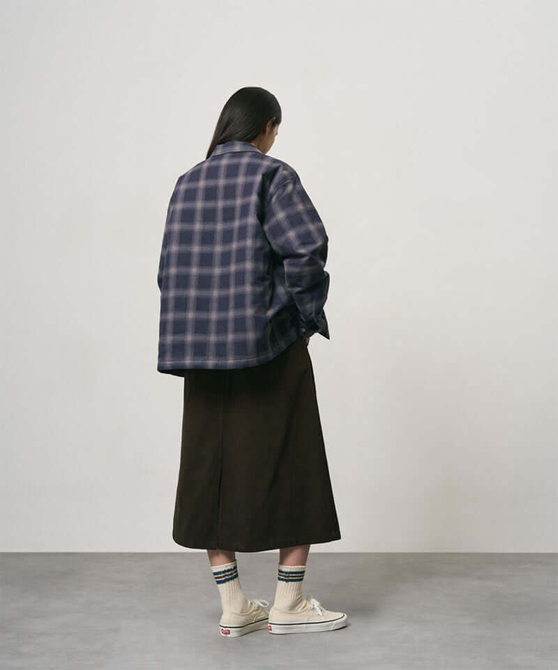 Back view of a model wearing a coffee colored Gramicci Venture Skirt and a blue plaid shirt on a light-tone background.