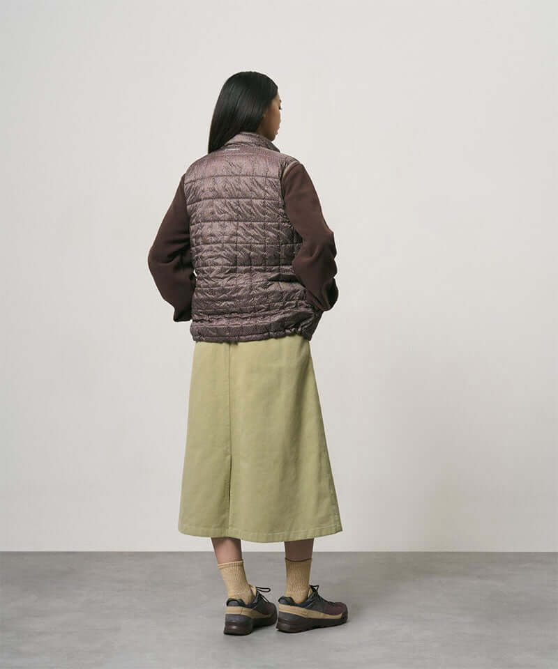 Back view of a model wearing a elm colored Gramicci Venture Skirt and a brown vest on a light-tone background.