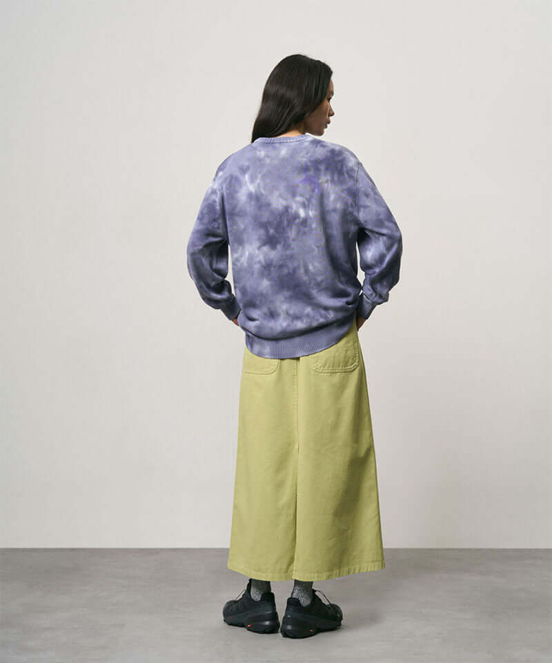 Back view of a model wearing a elm pigment colored Gramicci 5-Pocket Skirt and a purple tie-dye sweatshirt on a light-tone background.
