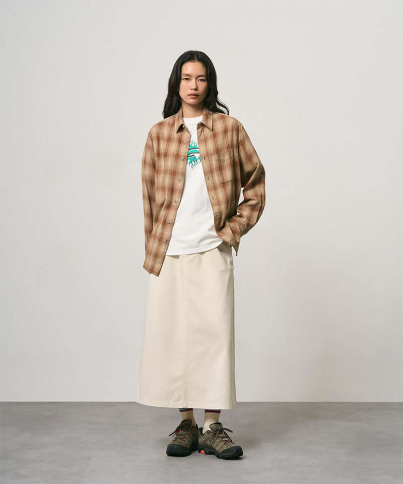 Model wearing a greige colored Gramicci 5-Pocket Skirt and a brown plaid shirt on a light-tone background.
