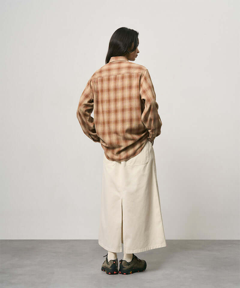 Back view of a model wearing a greige colored Gramicci 5-Pocket Skirt and a brown plaid shirt on a light-tone background.