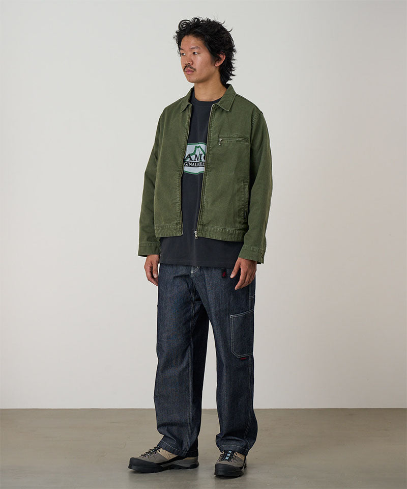 Model wearing a moss green Gramicci Taos Canvas Jacket and blue jeans on a light-tone background.