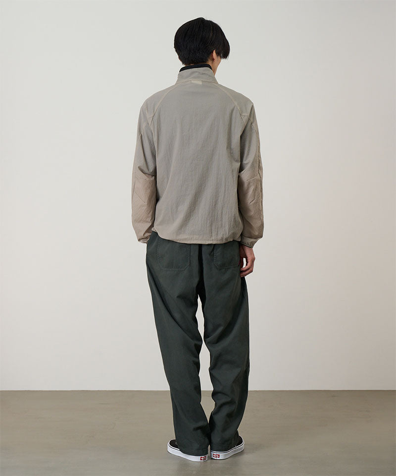 Back view of a model wearing a concrete colored Gramicci Ultra-Light Packable Jacket and dark grey pants on a light-tone background.