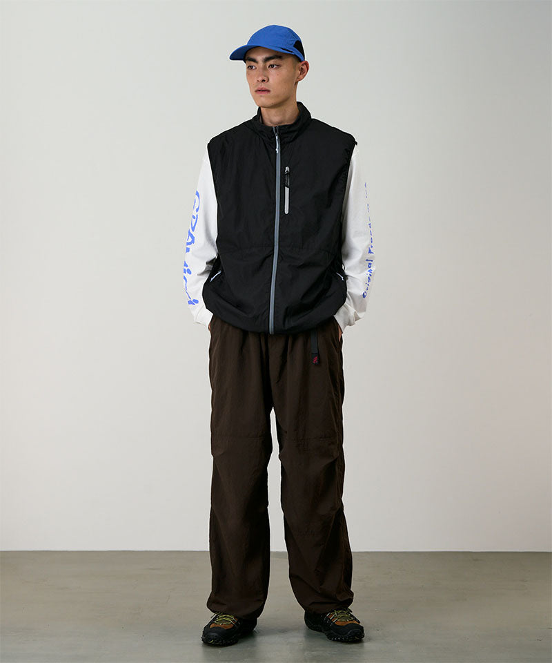 Person wearing a black vest, white shirt with blue text, brown pants, and blue cap against a plain background