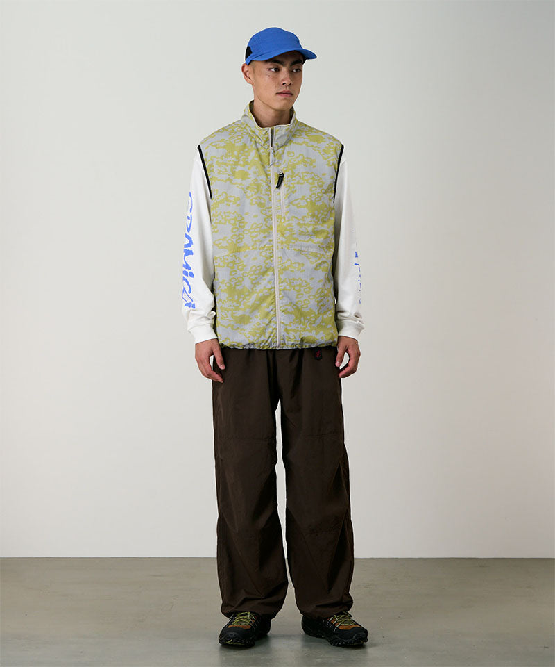 Person wearing a patterned vest, white long-sleeve shirt, brown pants, and blue cap against a plain background