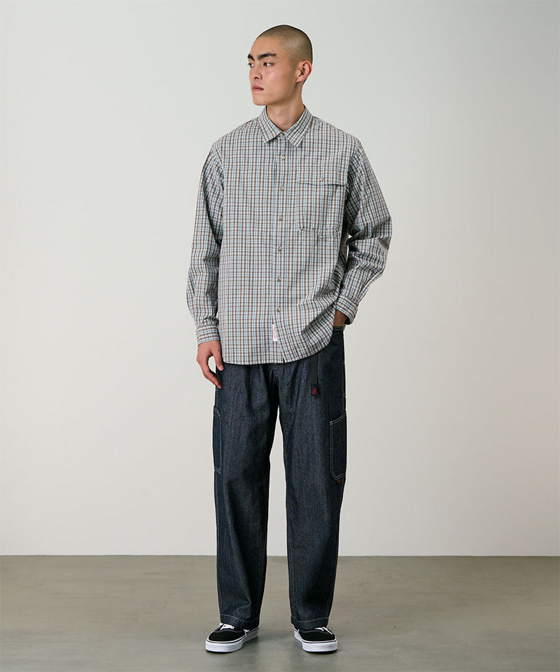 Model wearing a brown plaid Gramicci Poplin Millwood Shirt and blue pants on a light-tone background.