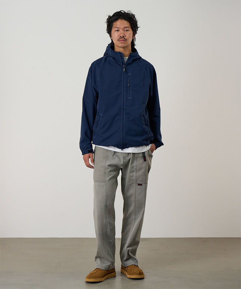 Model wearing a natural dyed olive Gramicci Gadget Pants and blue jacket on a light-tone background.