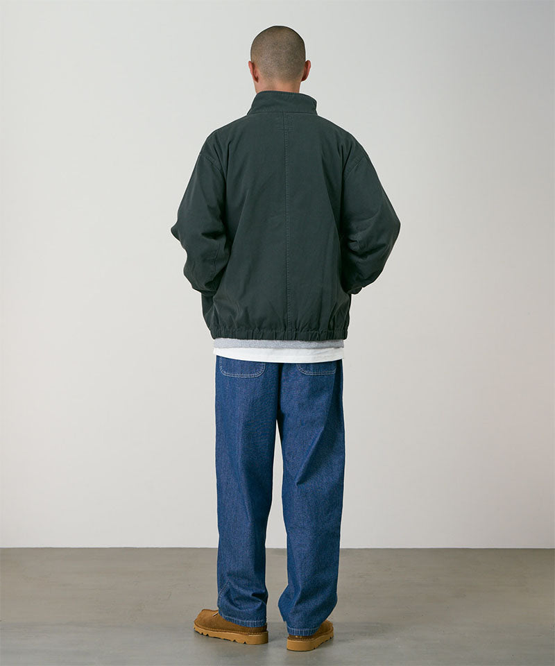 Rear view of a model wearing the Gramicci Japanese Chambray Double Knee pant in blue with a dark green jacket on a light tone background.