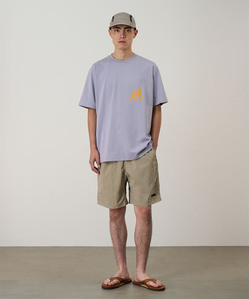 Man wearing a light purple t-shirt with a logo and beige shorts against a plain background