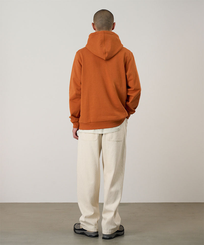Back view of a model wearing the Gramicci Blueberry Hooded Sweatshirt in orange with cream colored pants on a light tone background.