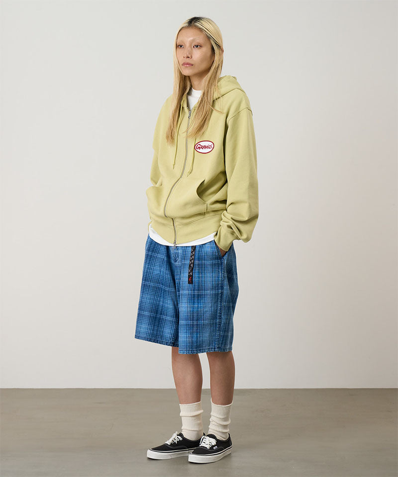 Person wearing a light green hoodie with a logo, white shirt, blue plaid skirt, and black sneakers on a plain background.