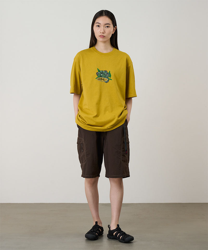 Model wearing a yellow Gramicci Blueberry Tee with a blueberry graphic and brown shorts on a light tone background.
