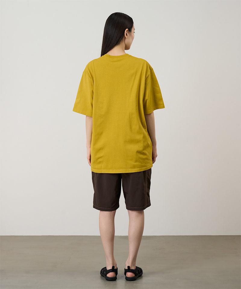 Back view of a model wearing a yellow Gramicci Blueberry Tee featuring a blueberry graphic with brown shorts on a light tone background.