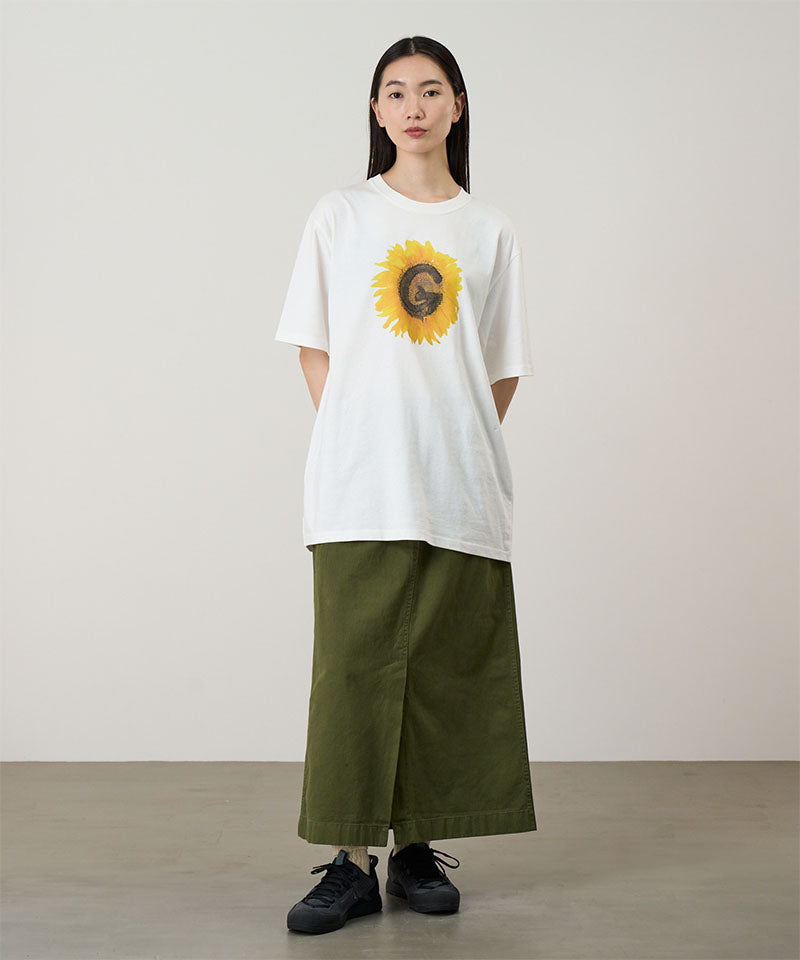 Model wearing a white Gramicci G-Flower Tee with a sunflower graphic and olive skirt on a light tone background.