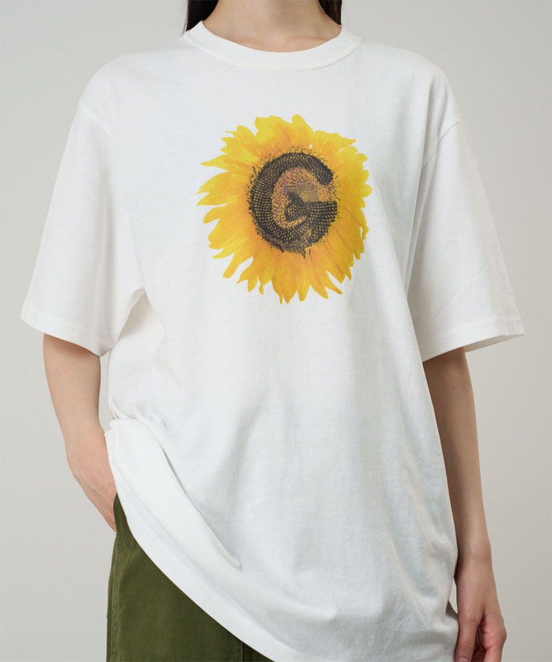 Model wearing a Gramicci white G-Flower Tee with sunflower graphic against a light tone background.