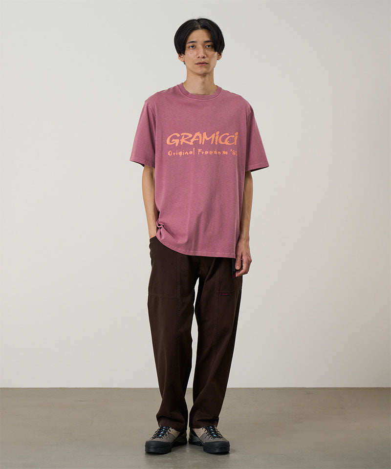 Model wearing a red pigment Gramicci Freedom Tee Pigment Dyed with a mango colored graphic and brown pants on a light tone background.