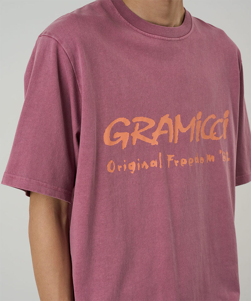 Model wearing a Gramicci red pigment Freedom Tee Pigment Dyed with mango colored graphic against a light tone background.