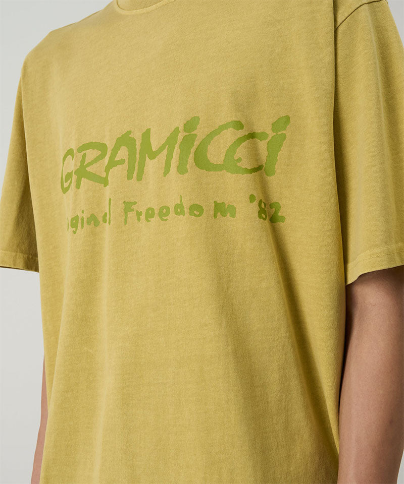 Model wearing a Gramicci yellow sulpher pigment Freedom Tee Pigment Dyed with lime green graphic against a light tone background.