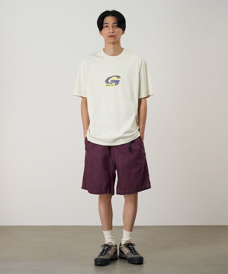Model wearing a natural colored Gramicci G-Scribble Tee Pigment Dyed shirt with a purple graphic and purple shorts on a light tone background.