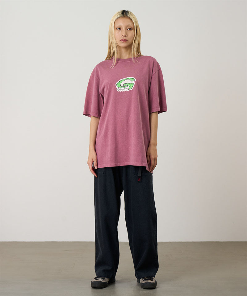 Model wearing a red Gramicci G-Scribble Tee Pigment Dyed shirt with a green graphic and green plaid shorts on a light tone background.