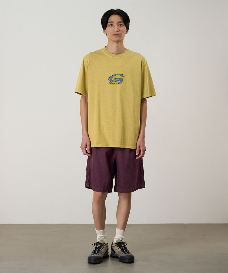 Model wearing a sulpher Gramicci G-Scribble Tee Pigment Dyed shirt with a blue graphic and purple shorts on a light tone background.